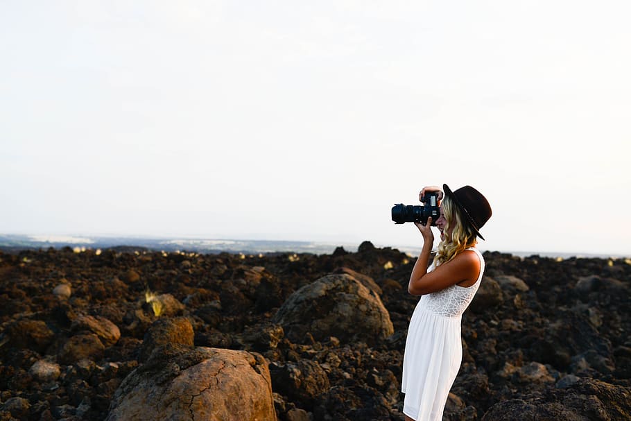 girl in white dress holding bridge camera while taking photo while standing on rock stone field at day time, woman holding camera HD wallpaper