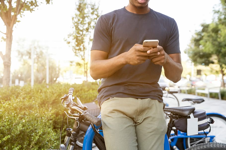 man holding smartphone leaning on bicycle during daytime, man wearing gray crew-neck t-shirt holding smartphone HD wallpaper