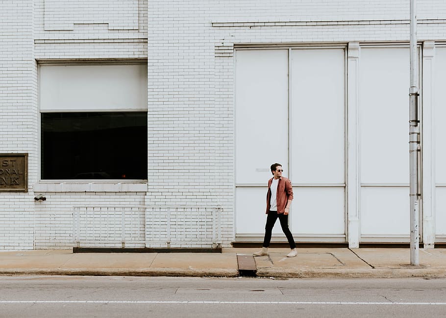 man walking on sidewalk beside white building during daytime, man wearing brown jacket walking near white steel gate