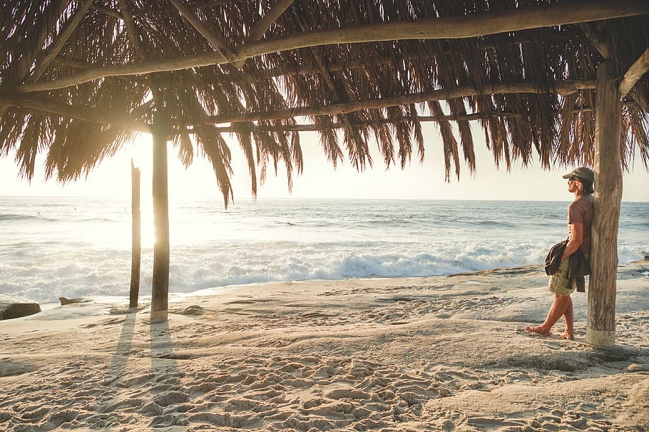 man leaning on wooden post while holding jacket, man leaning on hut post looking at beach HD wallpaper