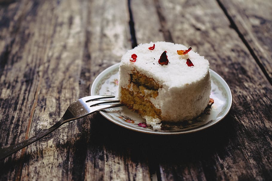 round icing-covered white cake on round white and green floral ceramic saucer beside gray fork on brown wooden table, round sliced cake on white ceramic plate beside gray stainless steel fork HD wallpaper
