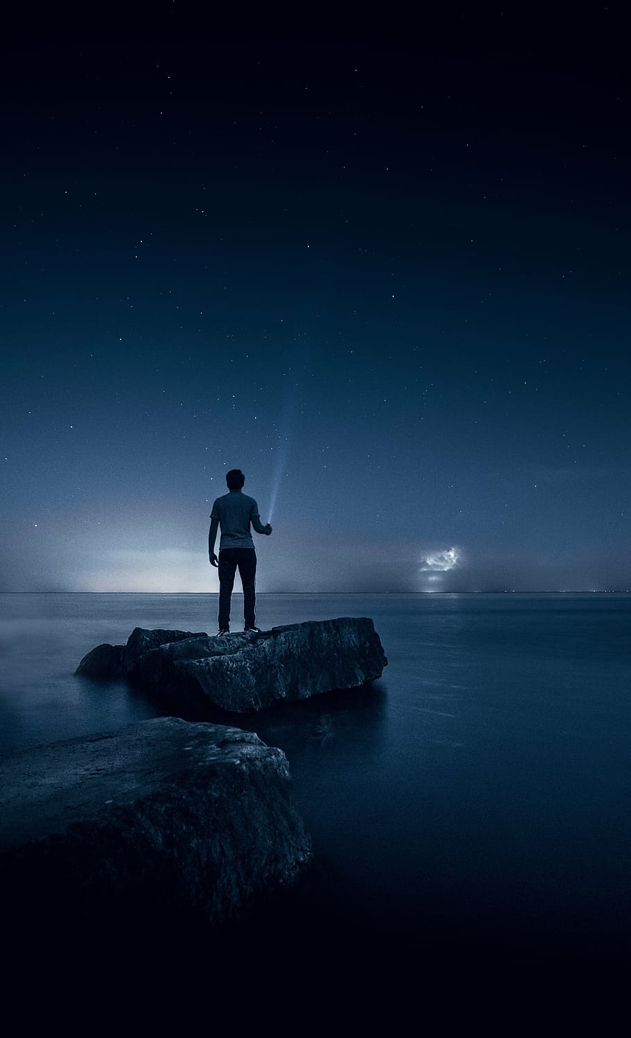 man standing on rock surrounded by water holding a flashlight pointing at the sky