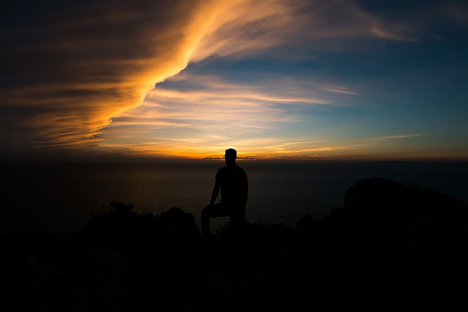 silhouette of person sitting on rock formation at golden hour, silhouette photo of man standing on peak surface during sunset