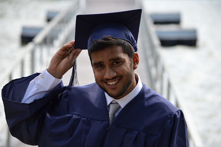 smiling man wearing academic dress stands beside railings at daytime, selective focus photography of man wearing blue academic gown closeup photo