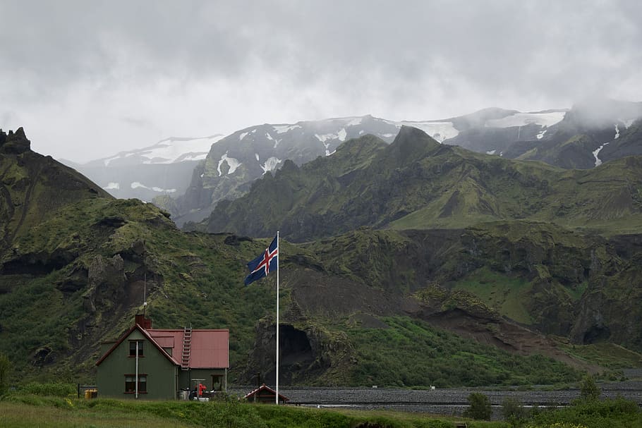 white concrete house with white, blue, and red cross flag near mountain range covered in green vegetation at daytime, blue and red cross flag with pole near gray house in front of river HD wallpaper