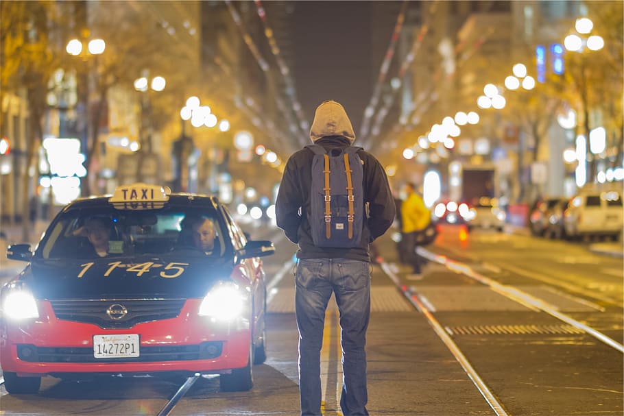 man wearing hoodie and backpack standing on middle of road, taxi