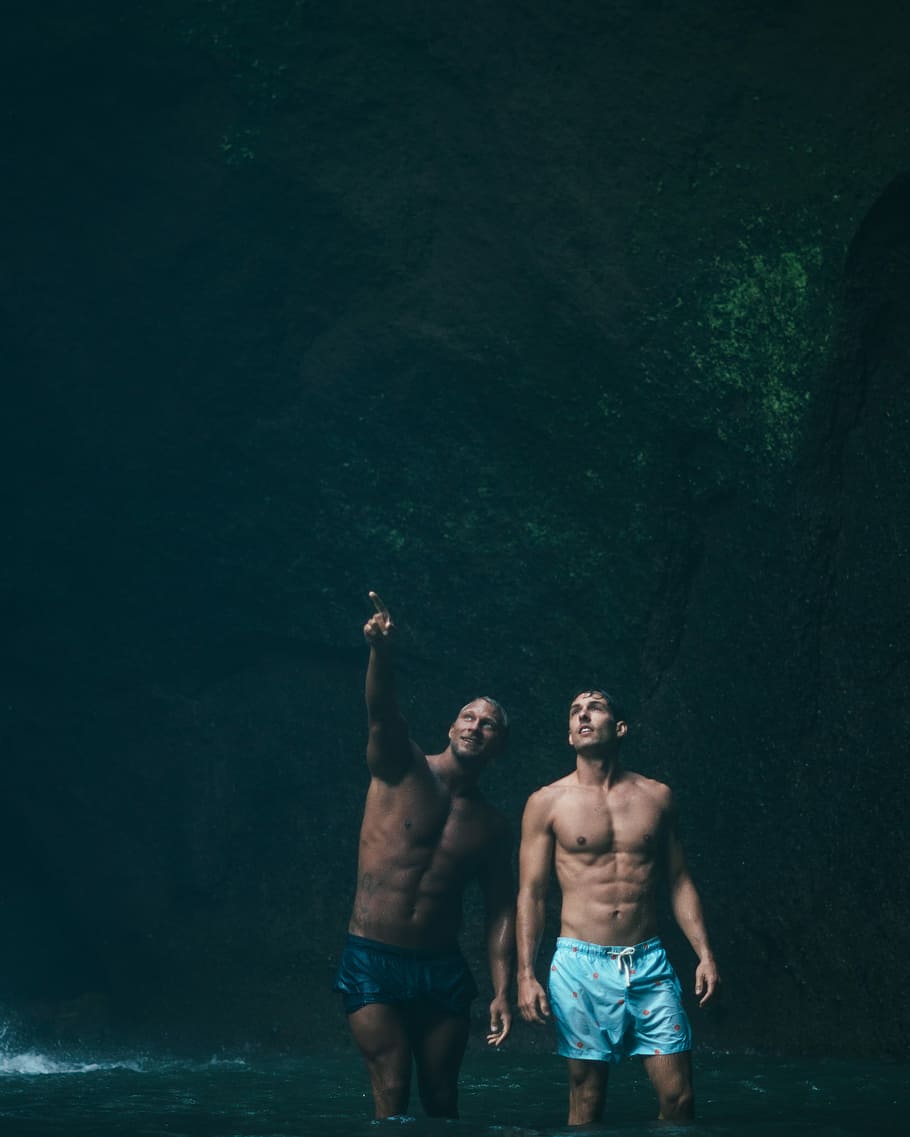 two men standing on body of water inside cave, two man in cave looking outside HD wallpaper