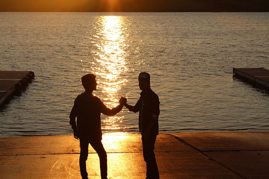 two standing man holding hands near ocean during sunset, best friends