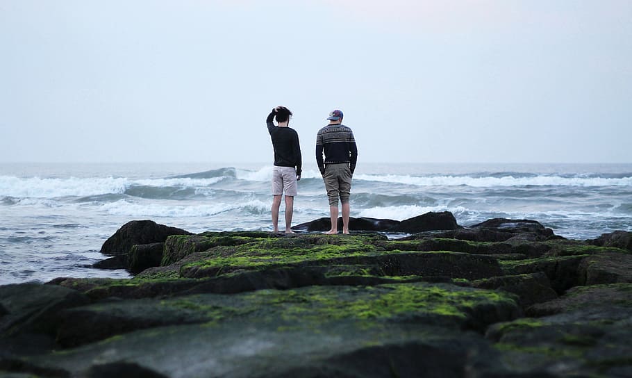 two man standing on rocks near sea during daytime, two men standing near body of water