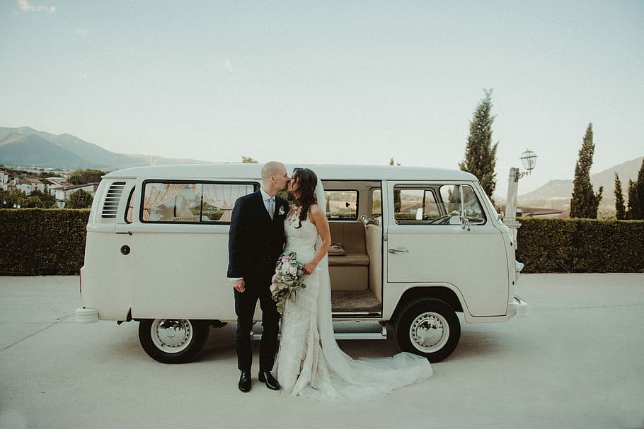 couple kissing beside white Volkswagen T2, woman in white wedding dress kissing man in black suit outdoors
