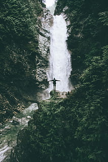 man standing at the cliff in front of the fall, person standing on rock formation in front of waterfalls during daytime HD wallpaper