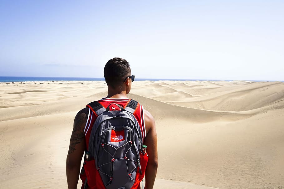 man looking at desert, man standing on brown sand wearing red tank top and black and gray backpack