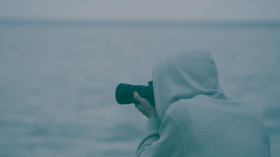 person wearing hoodie holding camera taking photo near body of water during daytime, person taking camera using DSLR camera during daytime, HD wallpaper