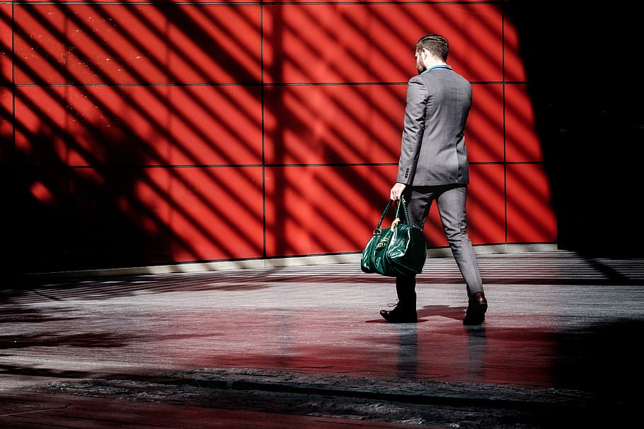 man holding green duffel bag while walking near red building at daytime, man walking near red concrete wall during daytime HD wallpaper
