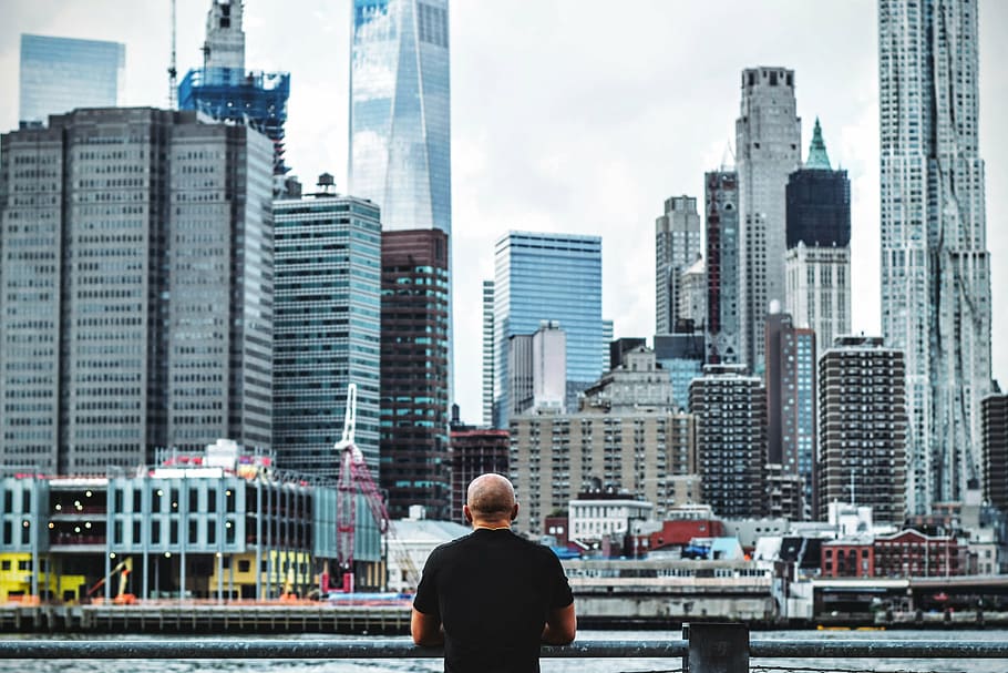 man wearing black shirt leaning on gray metal railing facing city buildings during daytime HD wallpaper