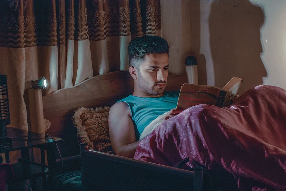 man in blue sleeveless top laying on bed reading book, man lying on the bed while reading book