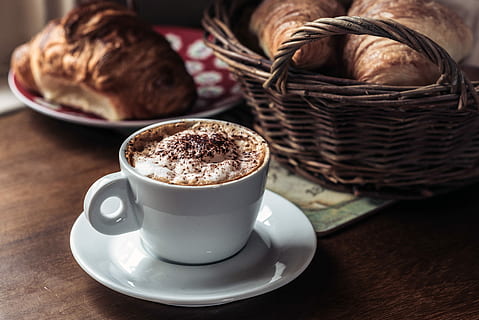 capuccino coffee on white ceramic cup with saucer beside pastry bread in basket and red plate, white ceramic teacup with liquid HD wallpaper