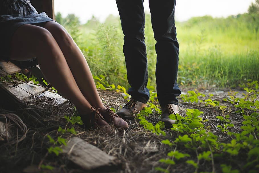 man and woman standing and sitting beside each other, person wearing blue jeans and brown shoes standing on brown soil, HD wallpaper