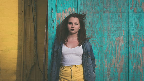 shallow focus photography of woman in white tank top and yellow jeans wearing blue denim jacket leaning on green wooden wall during daytime HD wallpaper