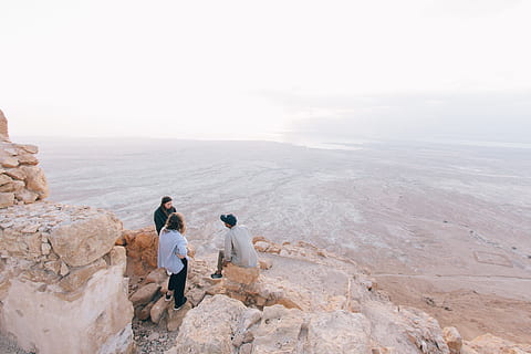 three person on cliff under white sky, three people standing and sitting on brown rock formation during daytime HD wallpaper