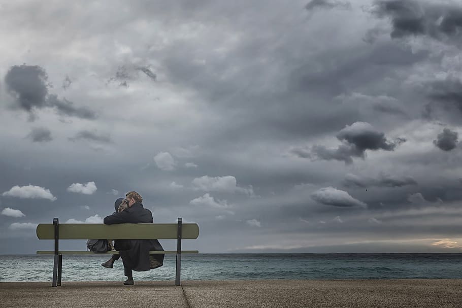 woman sitting on bench facing body of water under cloudy sky HD wallpaper