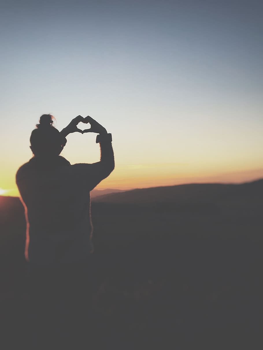 silhouette of a person making heart using her hands, photo of person doing heart hand sign facing mountain