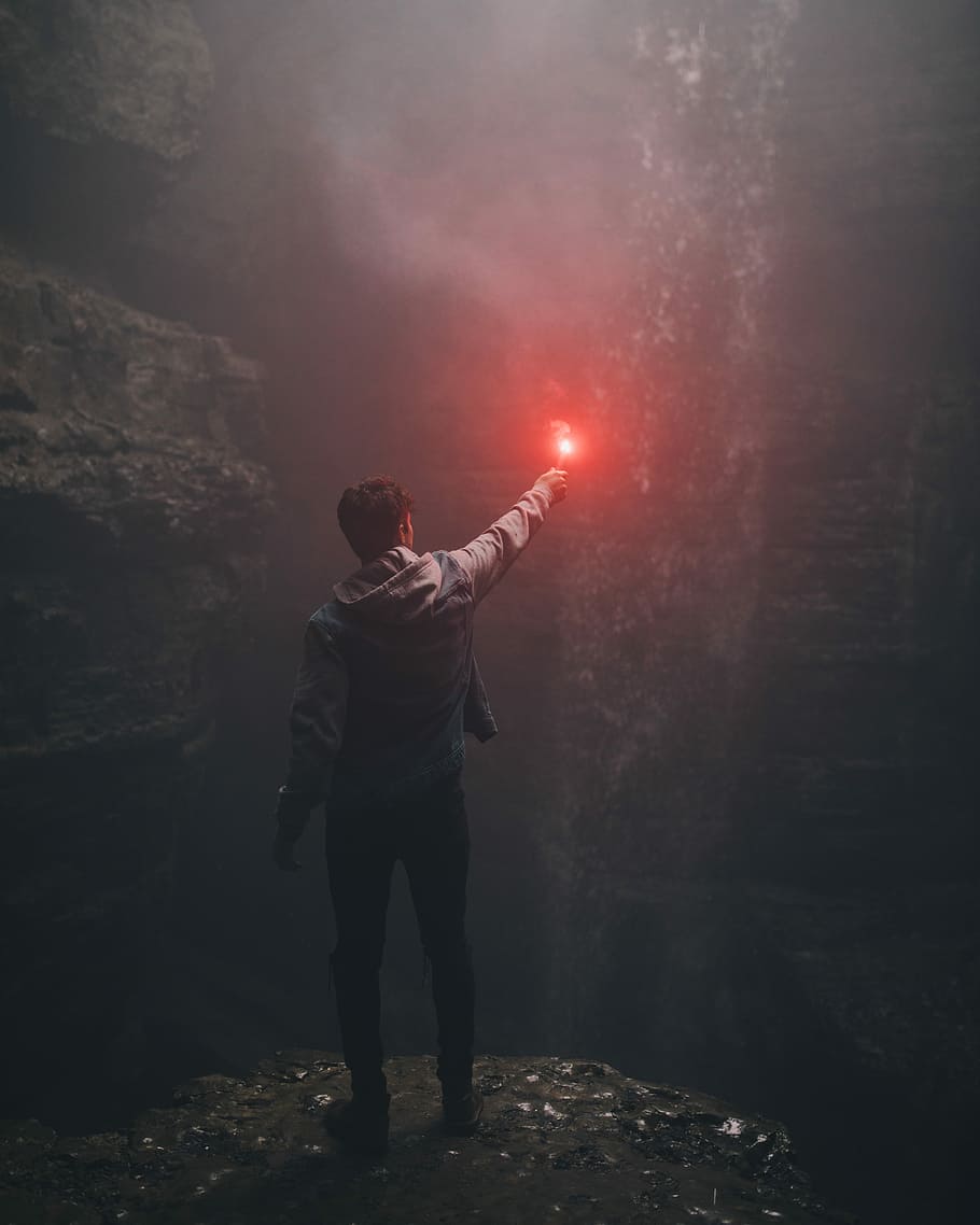 man lighting up flare in cave, man holding red light standing front of cave wall