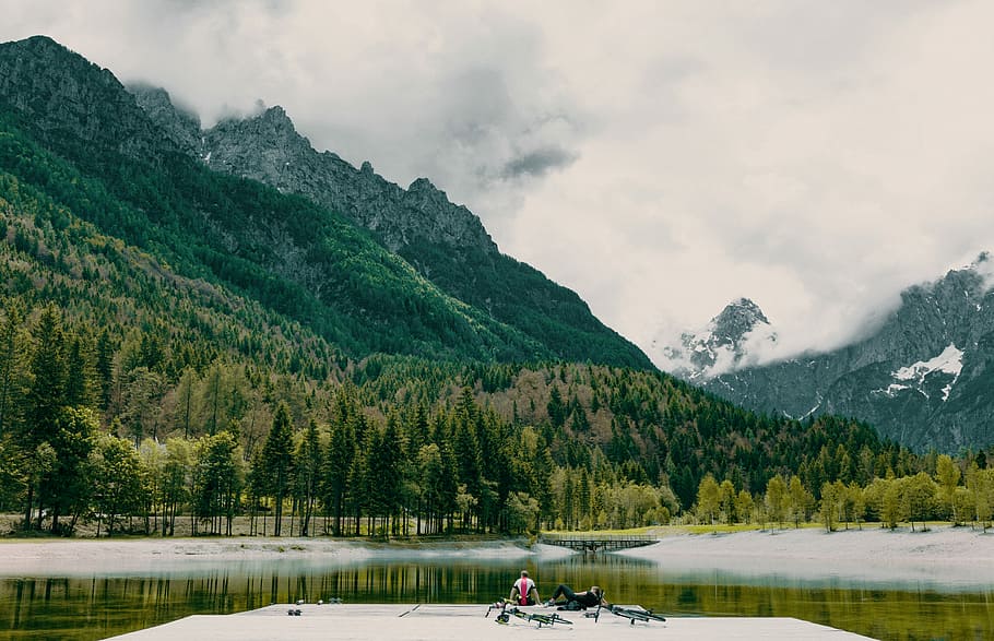 person staring at green body of water, two person sitting near body of water overlooking mountain range HD wallpaper