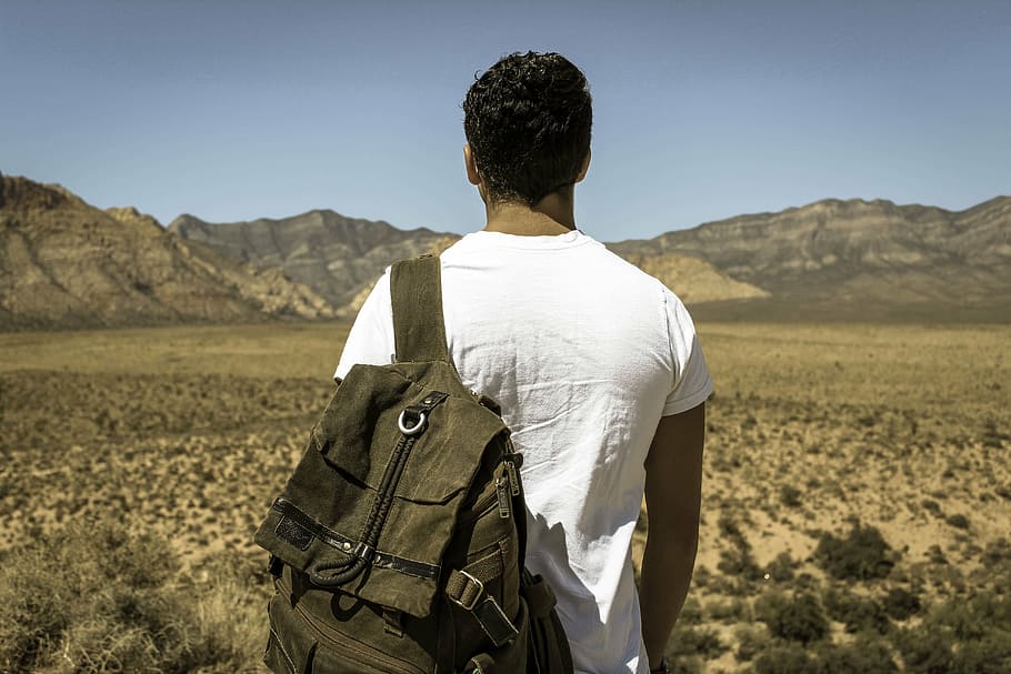 man in white shirt carrying backpack standing in front of mountain HD wallpaper