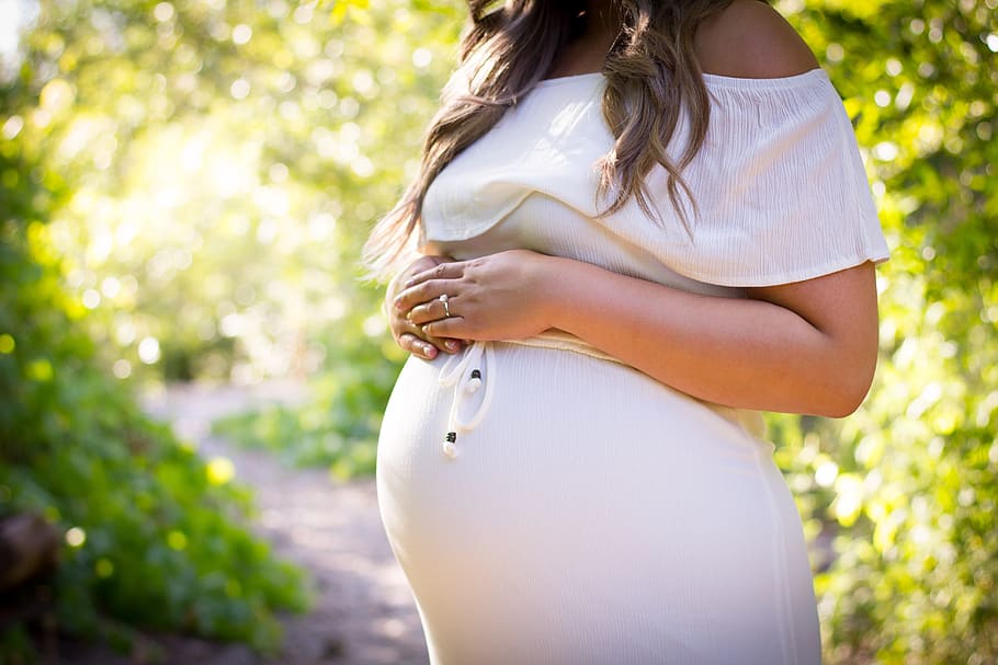 pregnant woman standing near green plants, woman wearing white off-shoulder maternity dress standing on pathway under green trees
