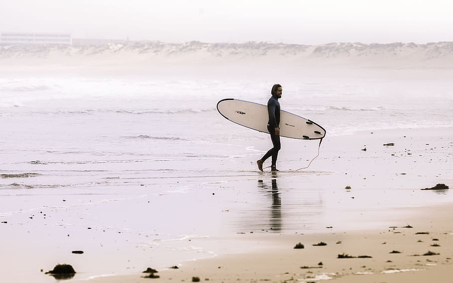 man holding surf board while standing on shore at daytime, man carrying white surfboard on seashore HD wallpaper