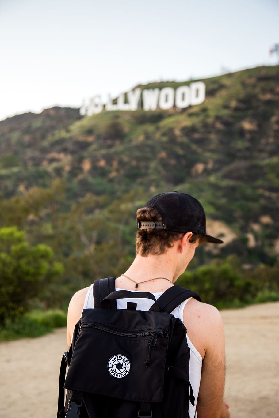 man wearing white tank top and black snapback cap carrying black backpack in distance hollywood signboard at daytime, selective focus photo of person wearing black backpack standing infront of Hollywood sign during daytime HD wallpaper