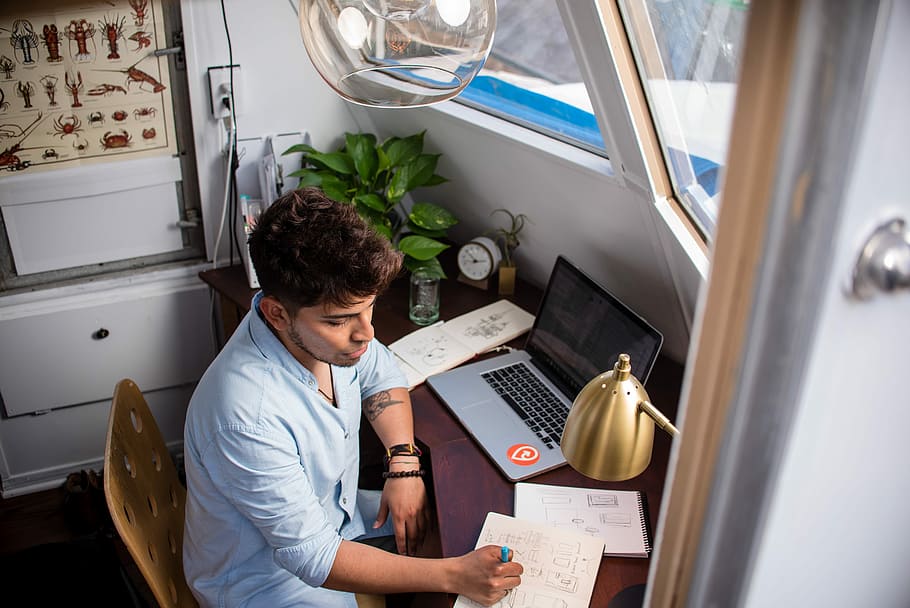 man sits while writing in front of MacBook, man writing on paper while sitting on chair during daytime HD wallpaper