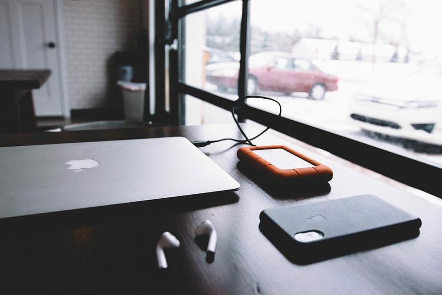 external hard drive plugged in MacBook near AirPods and iPhone X on table near window, iPhone X beside MacBook on brown wooden surface HD wallpaper