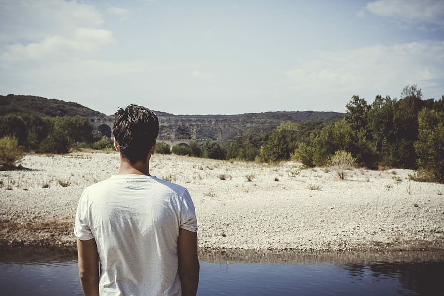 man standing on side of river, man in white crew-neck shirt looking at mountain HD wallpaper