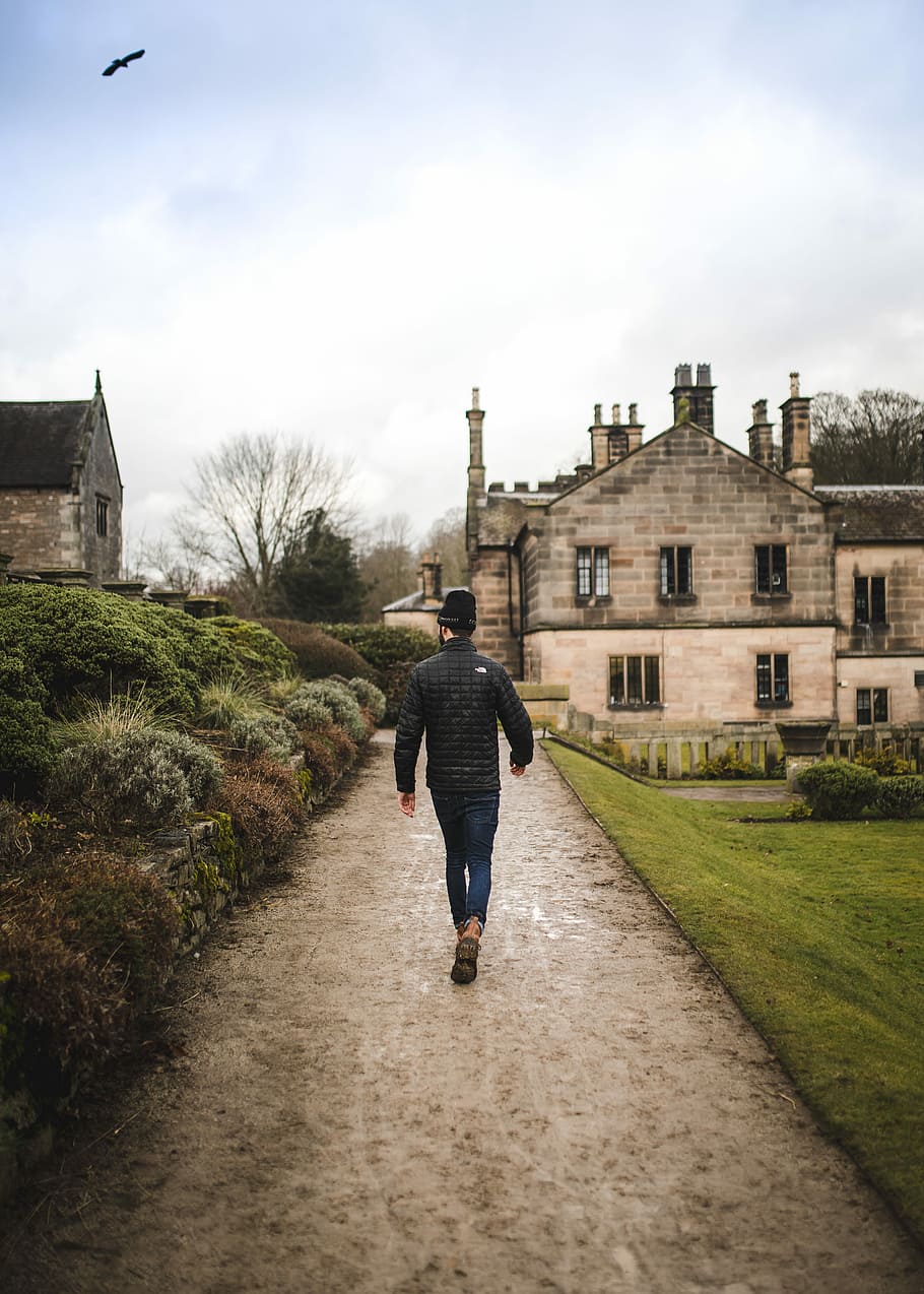 man walking on dirt road near brick house during daytime, man walking on pathway beside grass field, plants and buildings at the distance during daytime HD wallpaper