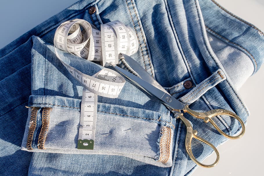 brown and grey garment shears on top of blue denim jeans, measuring tape