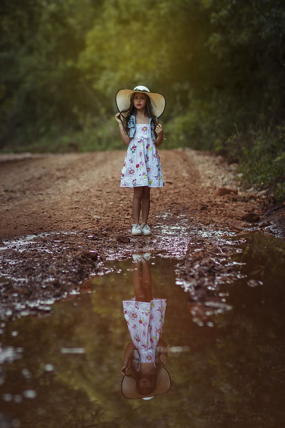 girl standing in front body of water, girl holding hair standing on dirt near road surrounded by trees during daytime
