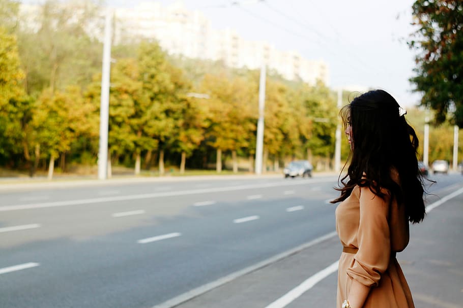 woman in brown dress standing by the road with car and green tress under grey clouds HD wallpaper