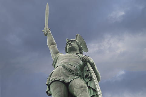 man raising sword and holding shield concrete statue under gray sky during daytime HD wallpaper