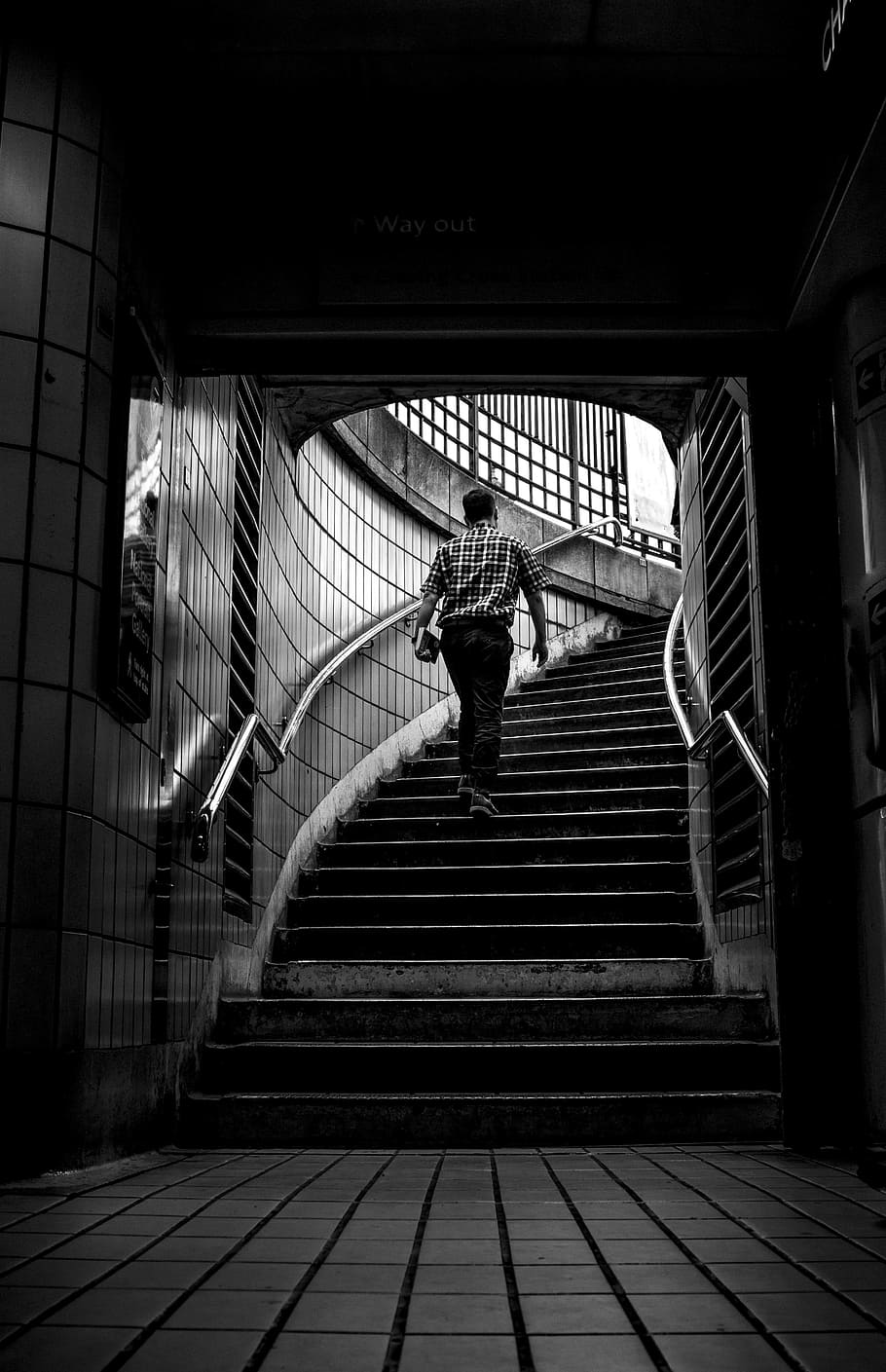 grayscale photo of man walking upstairs, grayscale photo of man carrying books while walking on stairs at a subway station