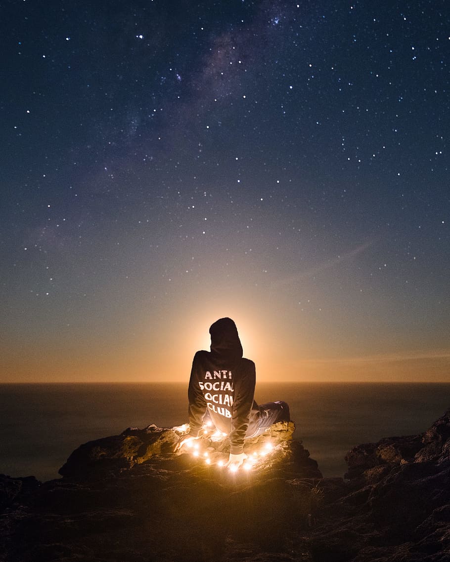 person wearing black anti social social club hoodie sitting on mountain cliff, person wearing blue Anti Social Social Club hoodie under blue starry night