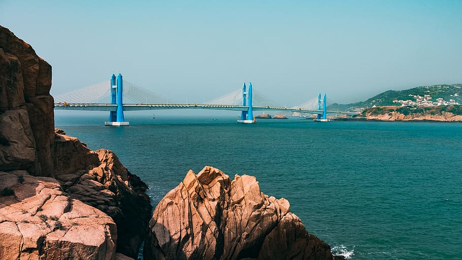 brown rock formation with blue and gray concrete bridge nearby above body of water, blue and white bridge under white sky, HD wallpaper