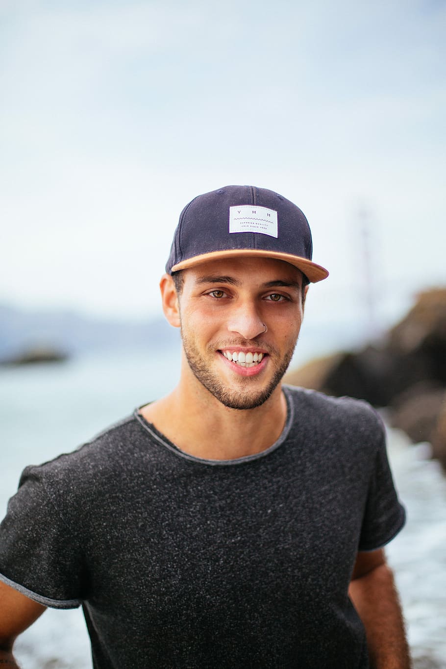 smiling man wearing black hat and black shirt at daytime, shallow focus photography of man in black crew-neck shirt showing smile HD wallpaper