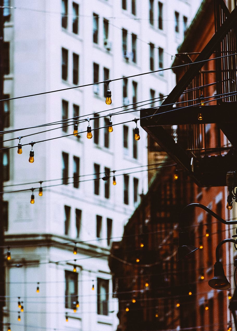 selective focus photography of string lights between concrete buildings, yellow light bulbs hanging on street HD wallpaper