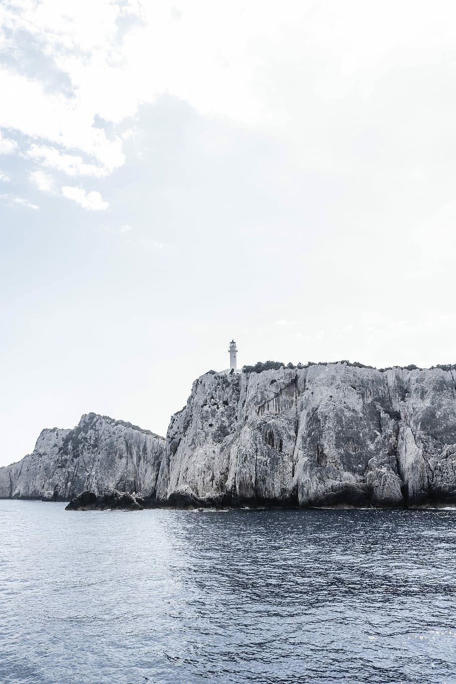 grayscale photography of lighthouse on cliff, white lighthouse at the cliff near body of water under white skies HD wallpaper