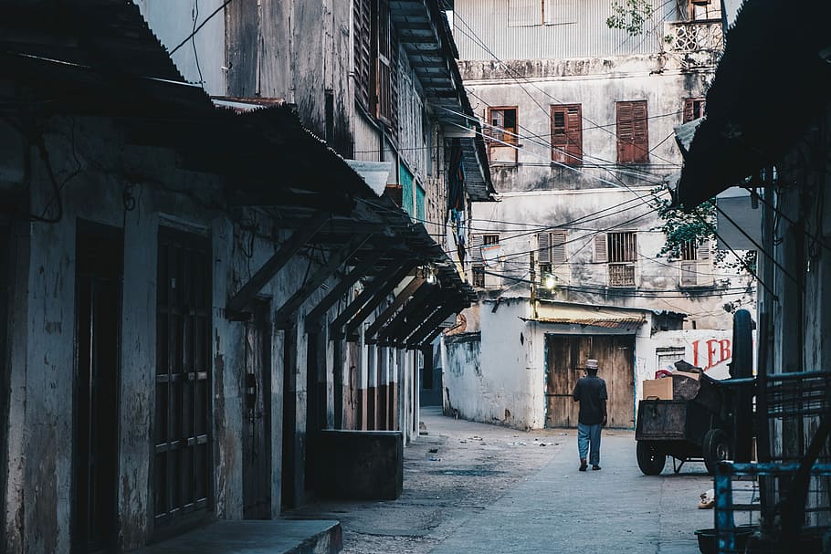 man walking near brown wooden cart between gray concrete buildings during daytime, man walking on street