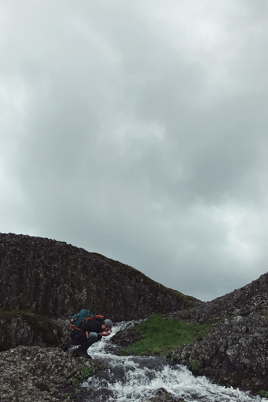 man kneeling near flowing water time lapse photo during cloudy day, mountain under grya sky HD wallpaper
