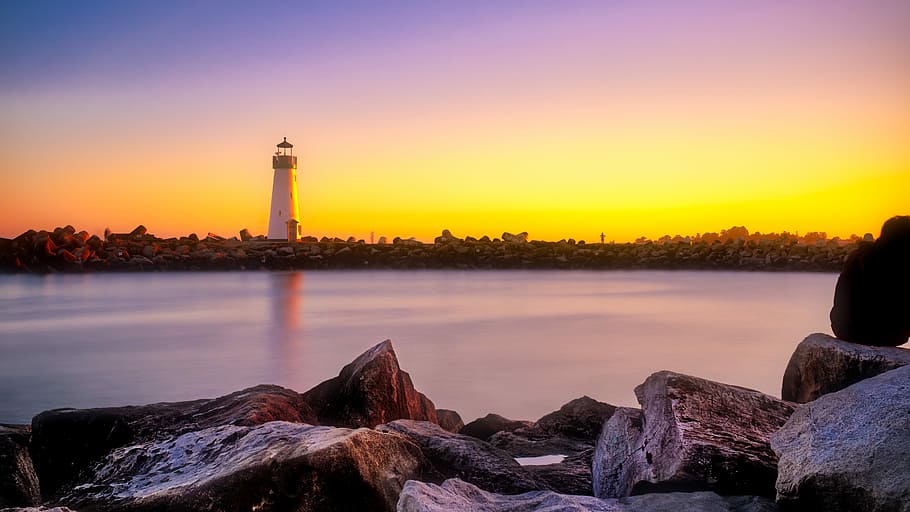 photo of rocks beside body of water with a view of lighthouse, white lighthouse near body of water during orange sunset HD wallpaper