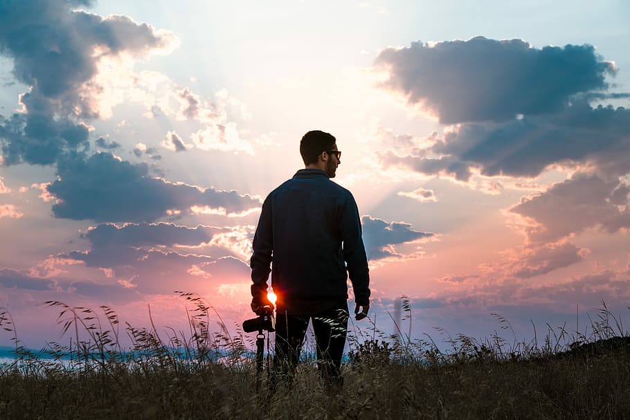 man in black long-sleeved shirt holding DSLR camera standing on grass, man standing on grass field holding DSLR camera HD wallpaper